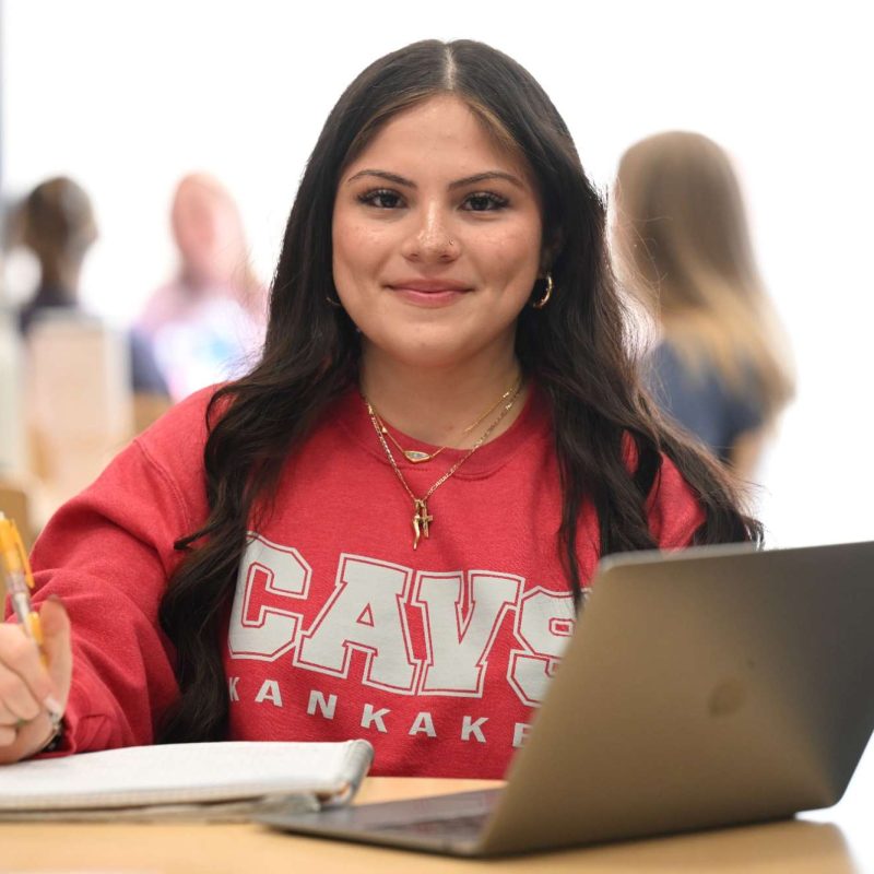 Student studying with a laptop and notebook, wearing a CAVS Kankakee sweatshirt, focused and smiling in a classroom setting.