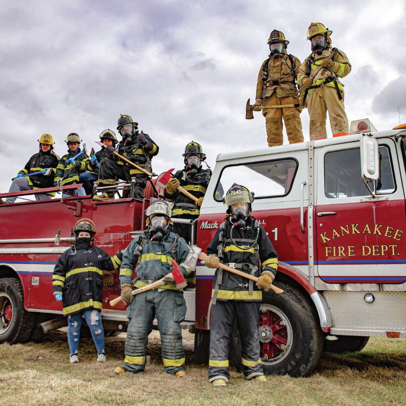 Firefighters from Kankakee Fire Dept. pose with gear and firetruck under a cloudy sky, showcasing teamwork and readiness.