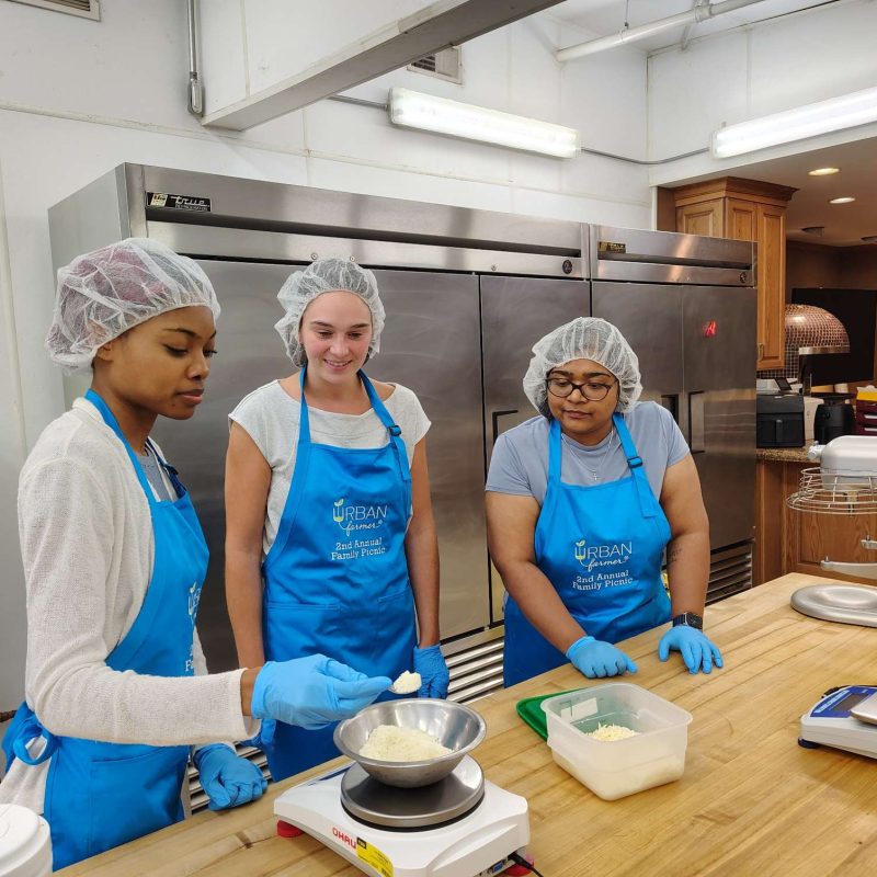 Three people in hairnets and aprons measuring ingredients in a kitchen, participating in Urban Recipes' cooking workshop.