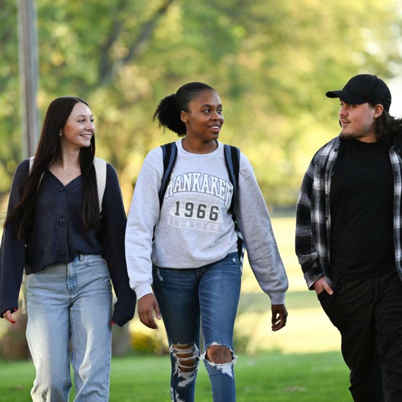 Three students walking together on a sunny campus lawn, engaged in conversation and smiling, Kankakee Community College.