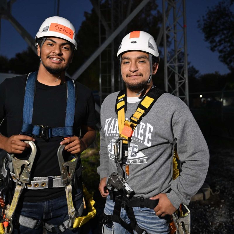 Two workers wearing safety harnesses stand outdoors, smiling, at Kankakee Community College.